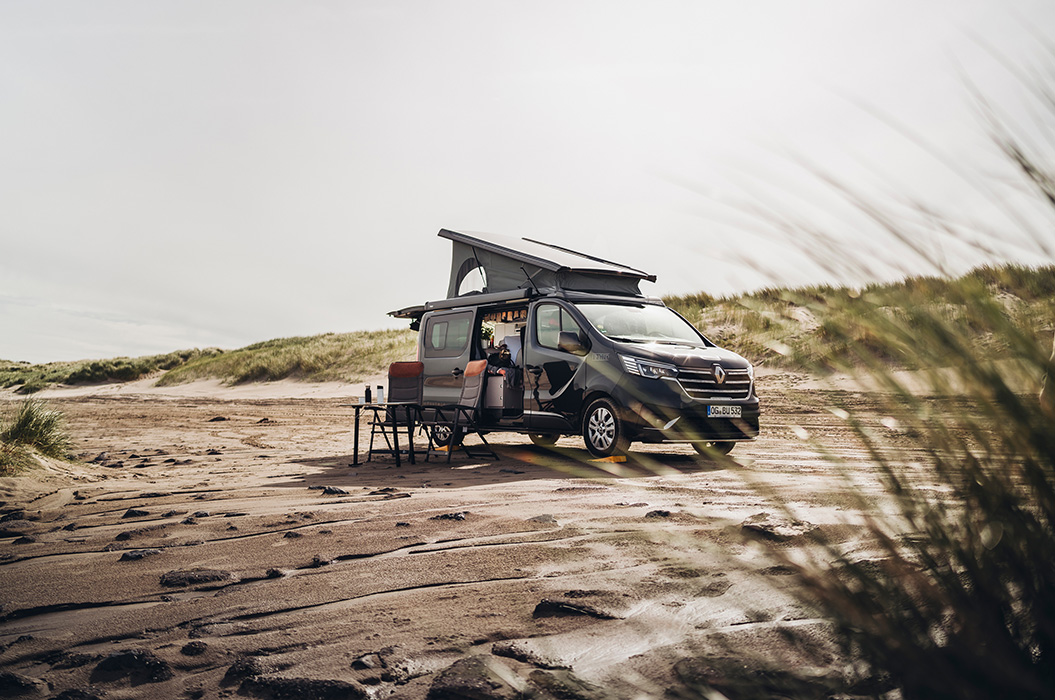 Ein grauer Camper mit Hubdach steht an einem sandigen Strand, Tisch und Stühle daneben.