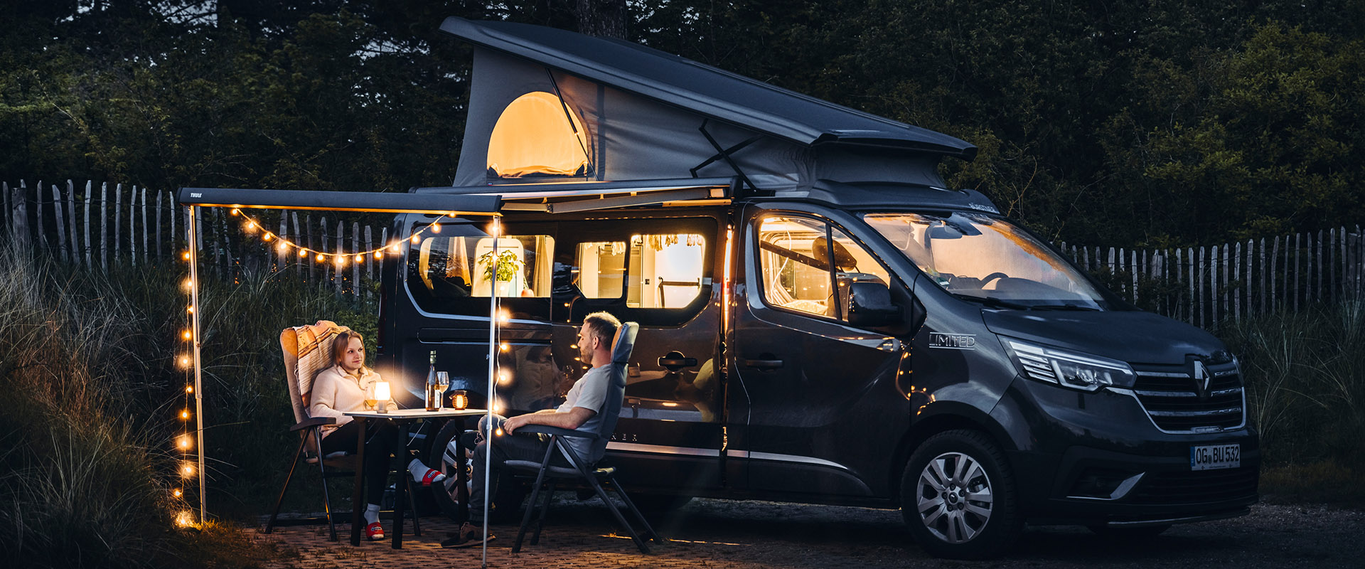 Zwei Personen, entspannt vor einem dunklen Camper mit Lichterkette und aufklappbarem Dach in der Natur.