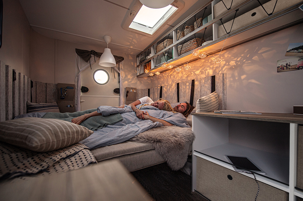 Paar entspannt im Bett, lächelnd. Wohnwagenambiente mit weichen Stoffen, warmes Licht, Kissen.