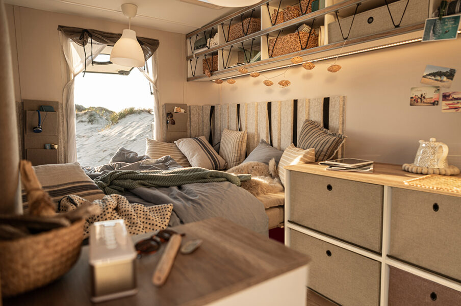Gemütliches Wohnmobilinterieur mit Strandblick, Bett mit Kissen und Decken, warme, neutrale Töne.