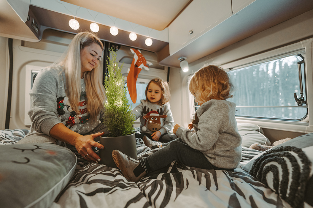 Eine Frau und zwei Kinder in einem Wohnmobil, alle lächeln und sitzen auf einem Bett mit Decken.