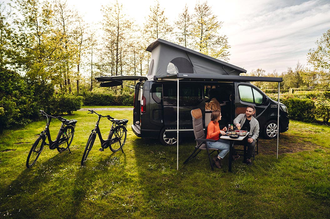 Drei Personen genießen entspannt ein Picknick neben einem schwarzen Camper mit Fahrrädern